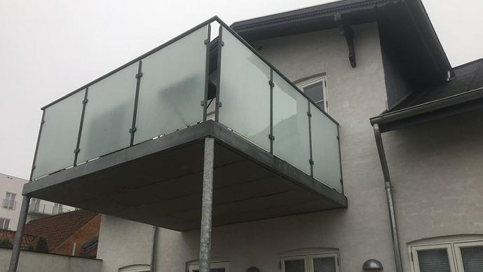 En moderne balkon med glasafskærmning, hævet over jorden på en hvid bygning.