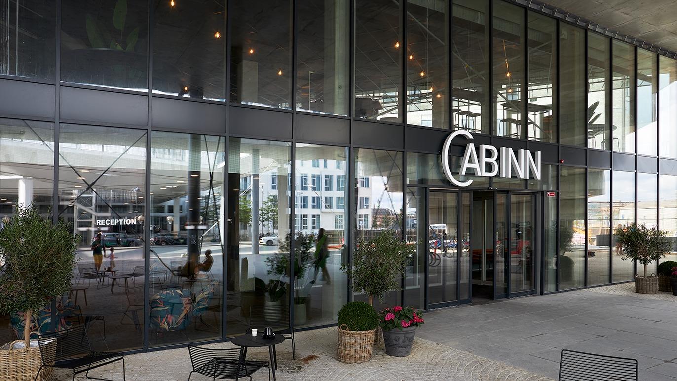 Moderne facade af CABINN hotel med store vinduer og indgang, omgivet af planter og udendørs siddepladser.