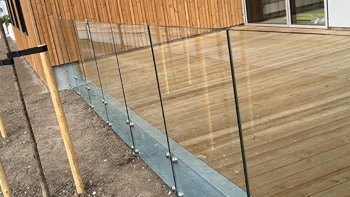 Glasrækværk på en træterrasse, der står på en betonfundament, med en moderne bygning i baggrunden.