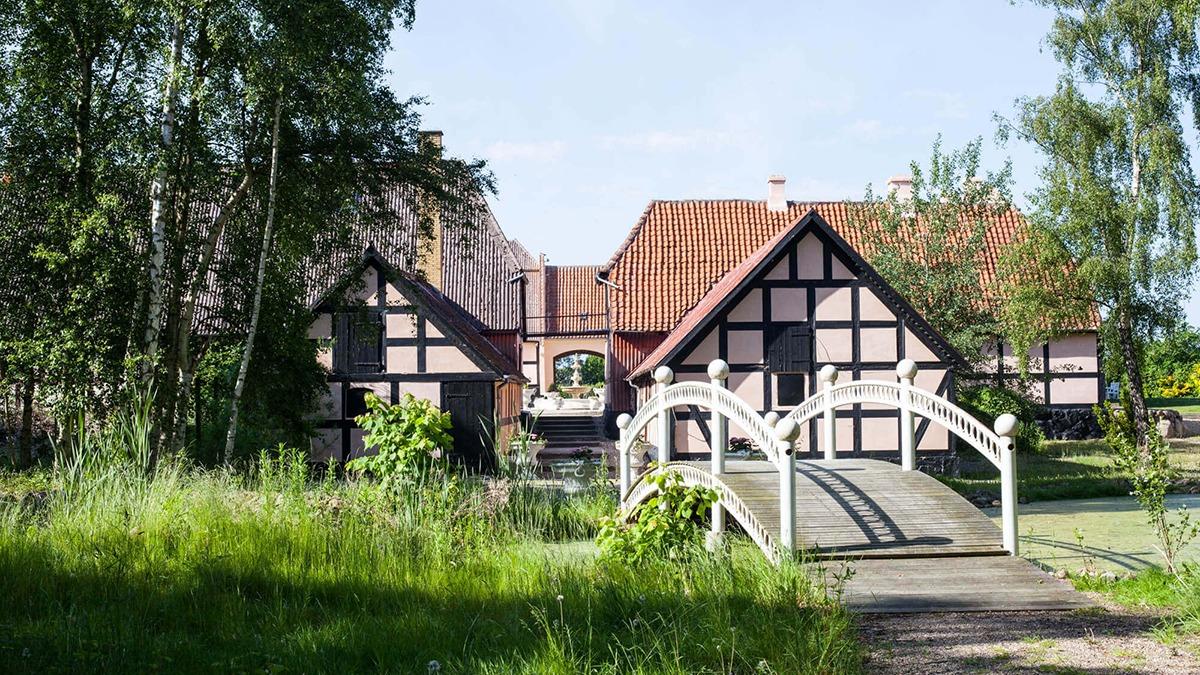 Et smukt bindingsværkshus med rød tag og en bro over en lille sø omgivet af grønne planter.
