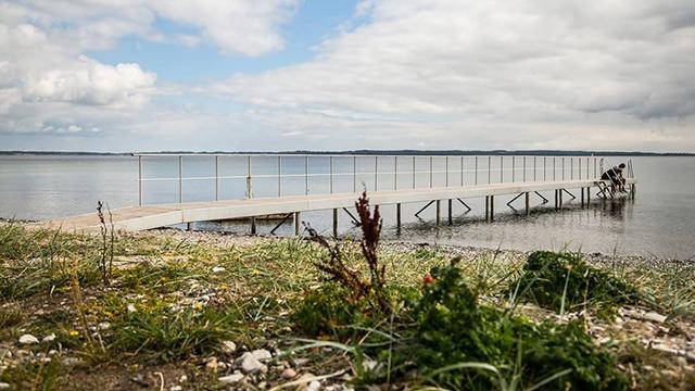 En træbro strækker sig ud over vandet med græs og planter i forgrunden under en skyet himmel.