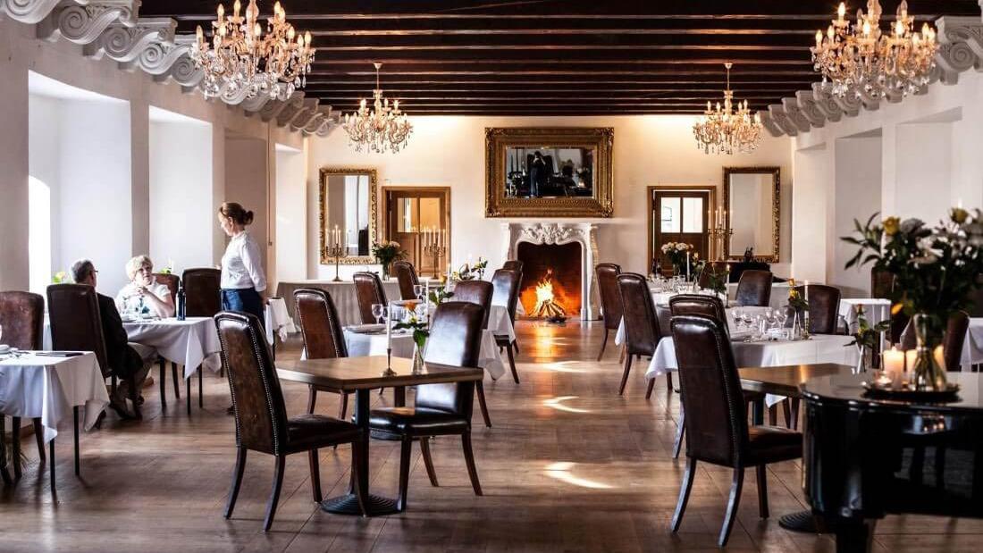 Elegant restaurant med lysekroner, borde dækket med hvide duge og en pejs i baggrunden.