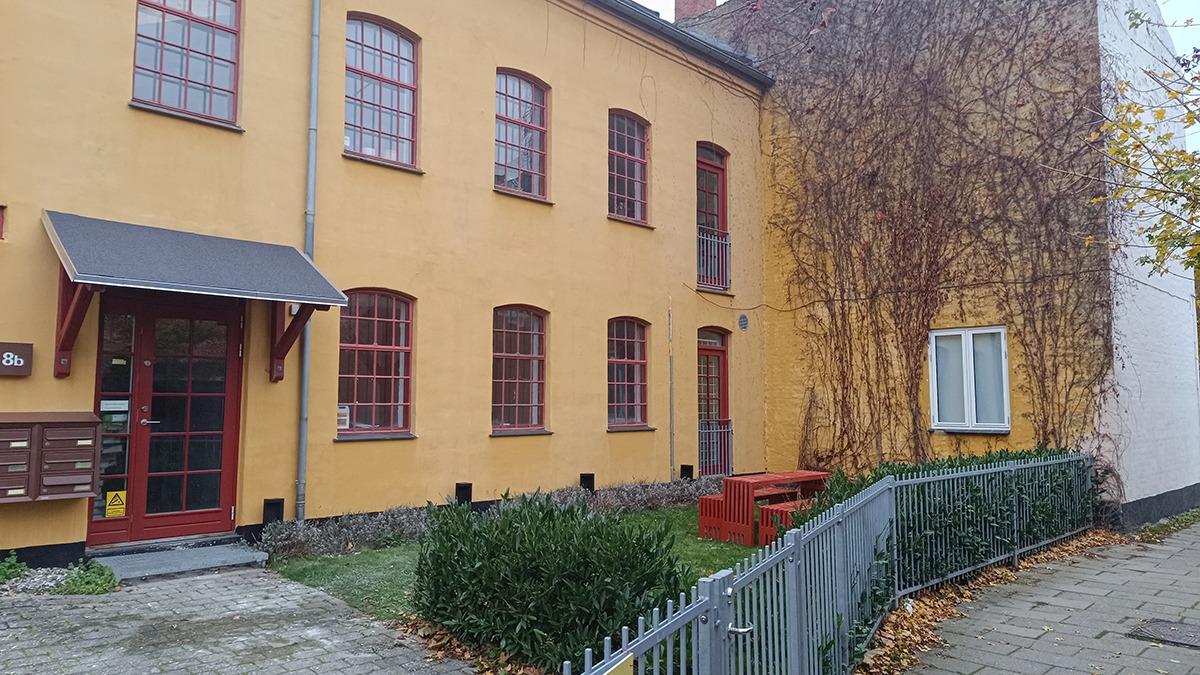 Gul bygning med røde vinduer og dør, grøn græsplæne og stakit i forhaven.