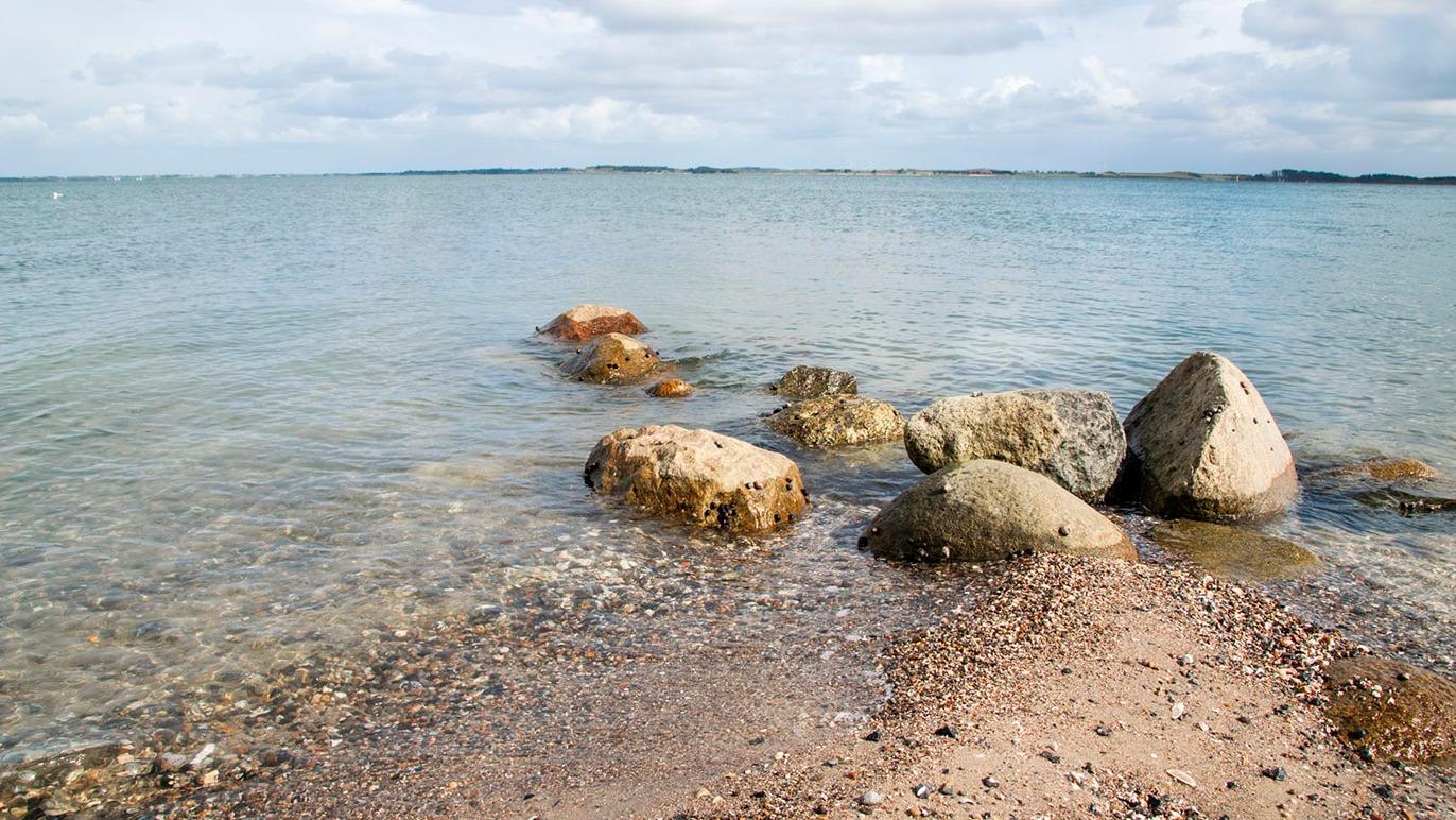 En rolig strand med store sten og klart vand, der strækker sig mod horisonten under en skyet himmel.