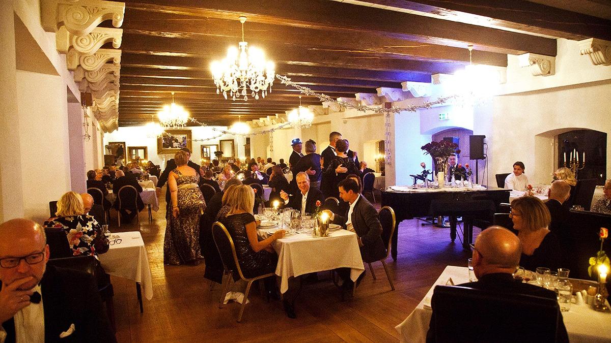En elegant restaurant med gæster, der spiser middag ved dækkede borde og levende lys, mens en pianist spiller.