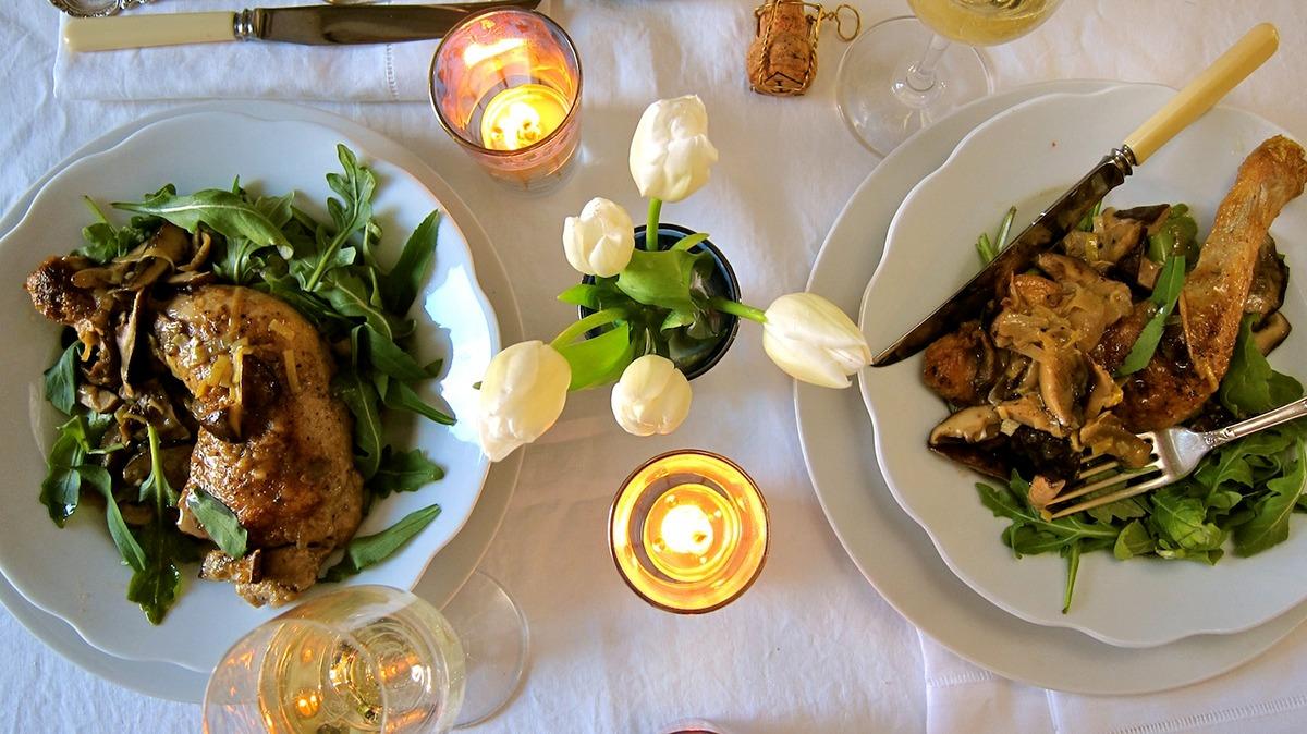 To plader med lækker mad, salat og stearinlys på et bord, omgivet af hvide tulipaner og vinglas.