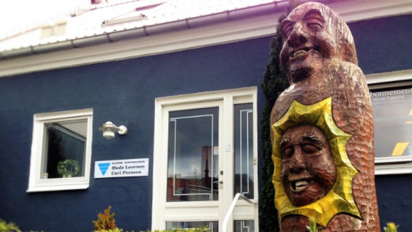 En farverig træskulptur af en sol og et ansigt foran en bygning med blå vægge og vinduer.