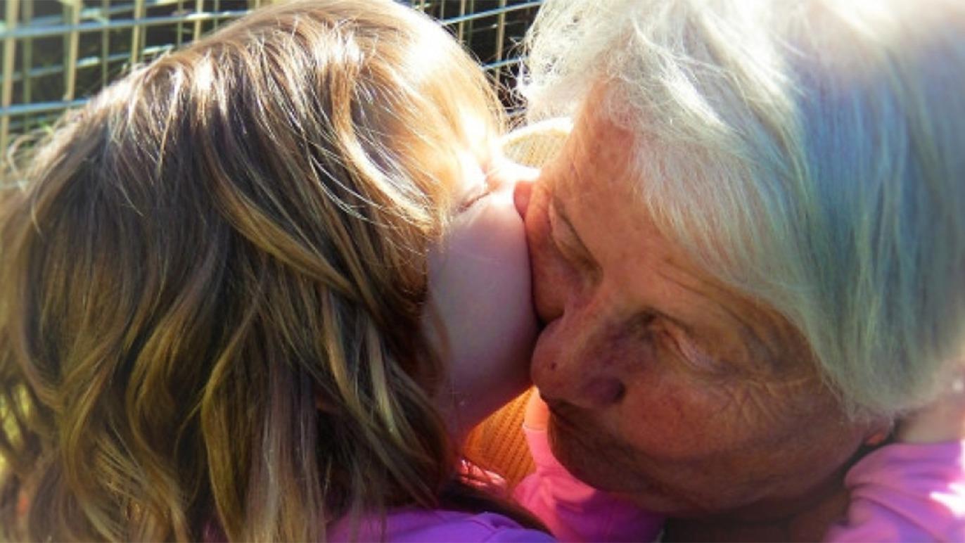Et barn kysser sin bedstemor i en solrig have, begge smilende og glade.