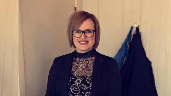 En kvinde med kort hår og briller iført en sort bluse med blomstermønster står smilende foran en væg.