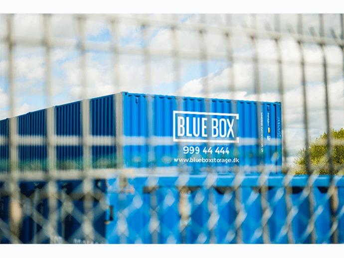 Blå container med teksten "BLUE BOX" og kontaktinformation, set gennem et hegn.