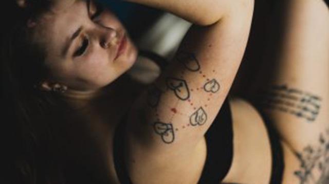 En ung kvinde med tatoveringer på armen, iført sort undertøj, sidder afslappet med hovedet bøjet.