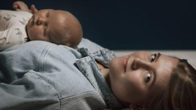 En mor ligger på en seng med sin nyfødte baby, der sover fredeligt på hendes bryst.