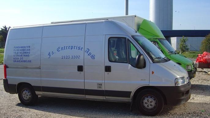 En sølvfarvet varevogn fra Enterprise med kontaktinformation på siden, parkeret ved en grøn lastbil.