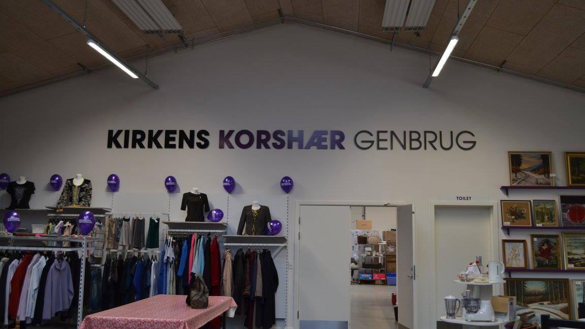 Butiksskilt for Kirken Korshær Genbrug med tøj og dekorationer i en genbrugsbutik.