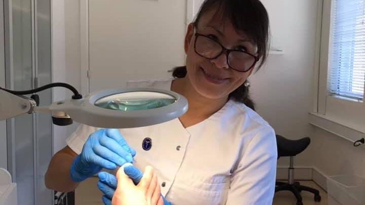 En smilende kvinde i hvidt arbejdstøj og blå handsker, der arbejder med en forstørrelseslampe.