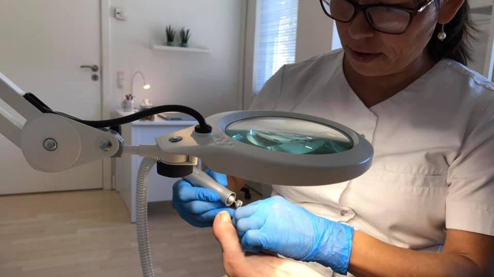 En kvinde i hvidt arbejdstøj udfører en neglebehandling under et forstørrelsesglas.