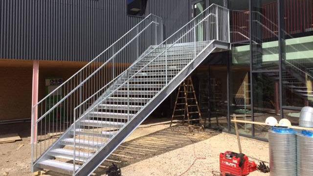 En metaltrappe med gelænder fører op til en bygning, omgivet af byggeudstyr og en stige.