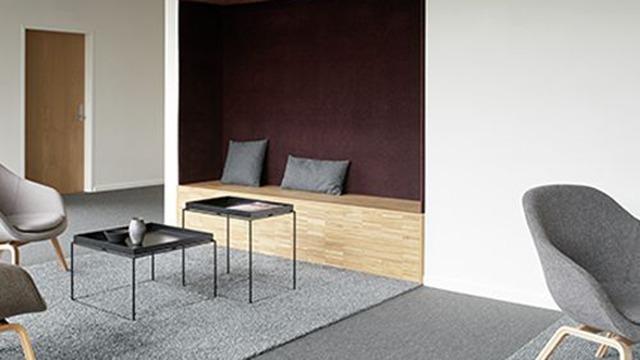 Moderne indendørs lounge med grå stole, sorte borde og en træbænk med puder på en grå tæppe.