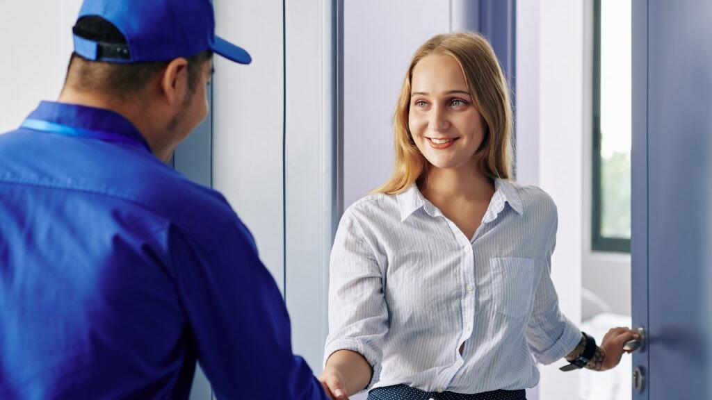 En kvinde åbner døren og hilser på en mand i uniform, der står med et smil.