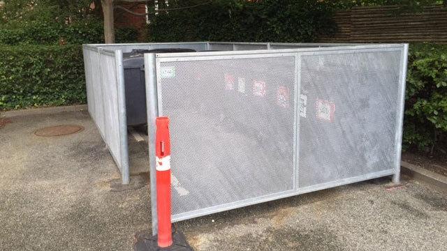 Affaldscontainer indhegnet med metalgitter og rød sikkerhedspæl ved en parkeringsplads.