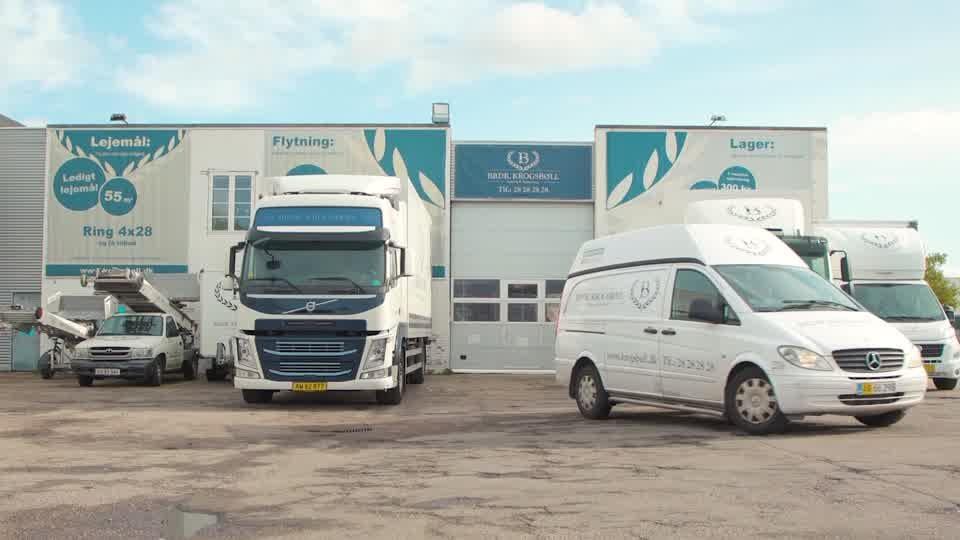 Lagerbygning med lastbiler og varevogne parkeret foran, med logoer og skiltning synligt.