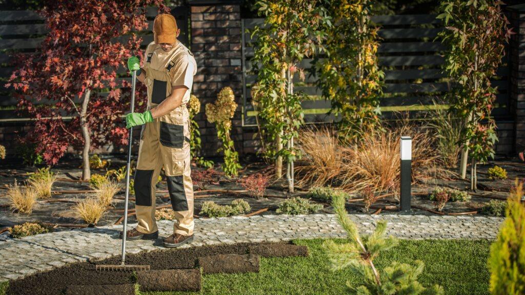 En gartner arbejder i haven med en rive, omgivet af planter og dekorative buske.