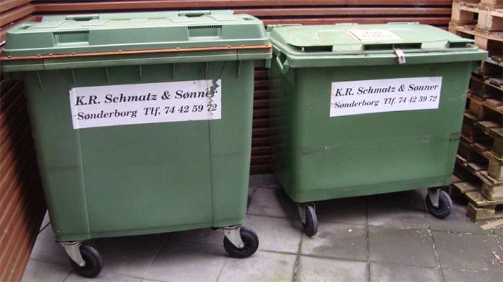 To affaldscontainere fra K.R. Schmidt & Sønne i Sønderborg, placeret ved en væg med træpaller i baggrunden.