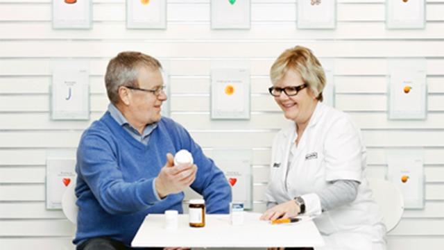 Ældre mand og kvinde i hvidt arbejdstøj sidder ved bord og diskuterer medicinflaske og piller.