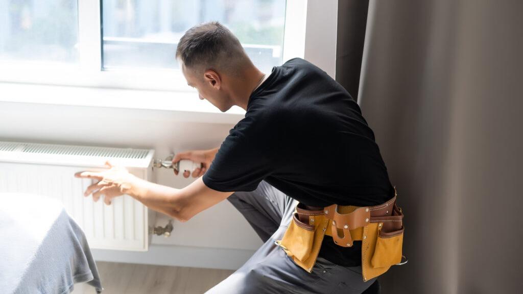 En håndværker arbejder på en radiator i et lyst rum med en værktøjsbælte om livet.