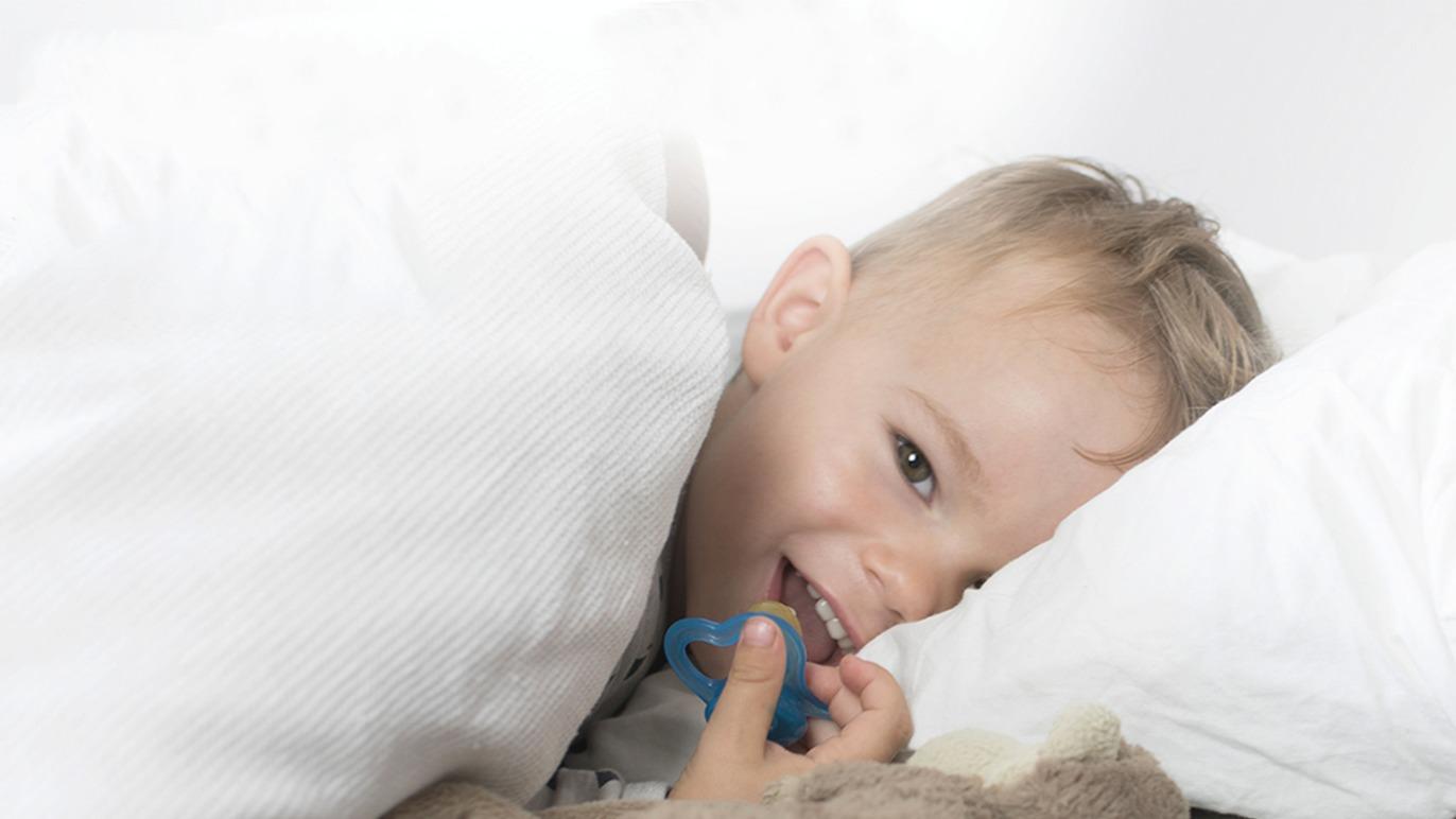 Lille dreng ligger under tæppet med sut og smiler, omgivet af puder og en bamse.