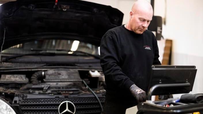 Mekaniker arbejder på en Mercedes bil i et værksted med en computer ved siden af.