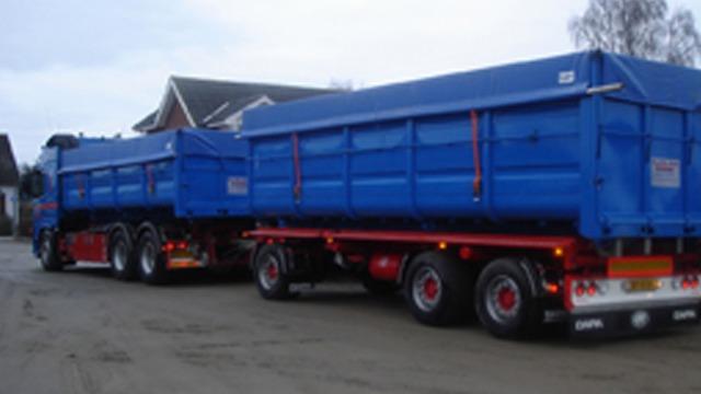 Blå lastbil med tilhørende trailer parkeret på en vej ved bygninger.