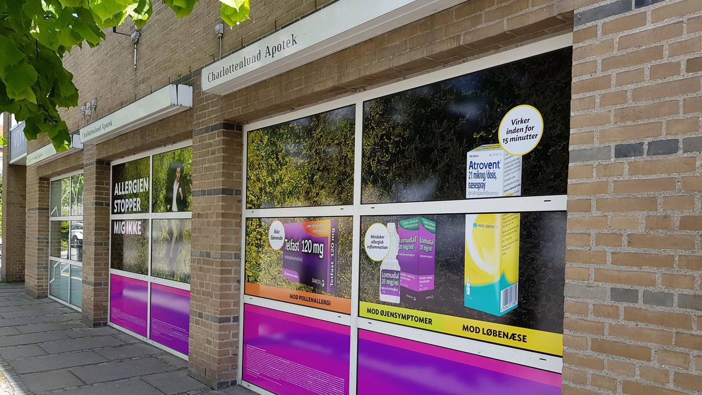 Butiksfacade med store vinduer, der viser farverige reklamer for forskellige produkter og tjenester.
