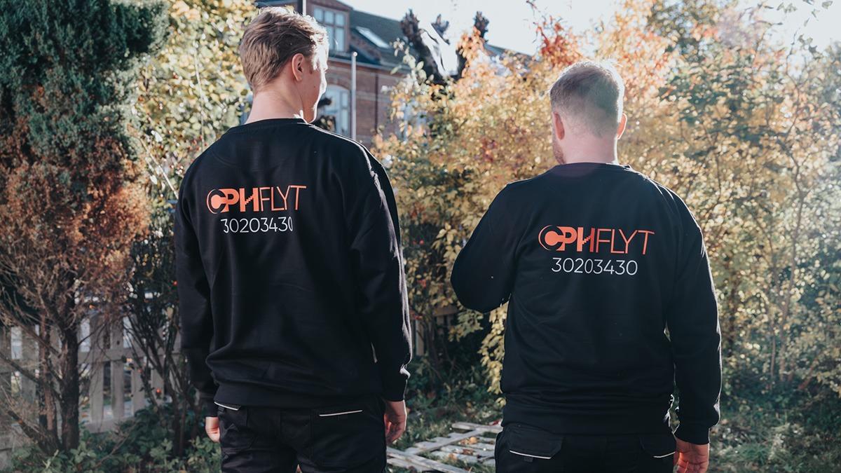 To personer iført sorte sweatshirts med logoet "OPHFYLT" på ryggen, går gennem en have med efterårsfarver.