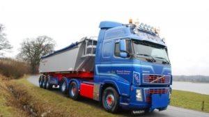 Blå lastbil fra Volvo med en grå trailer, der kører langs en vej ved en sø.