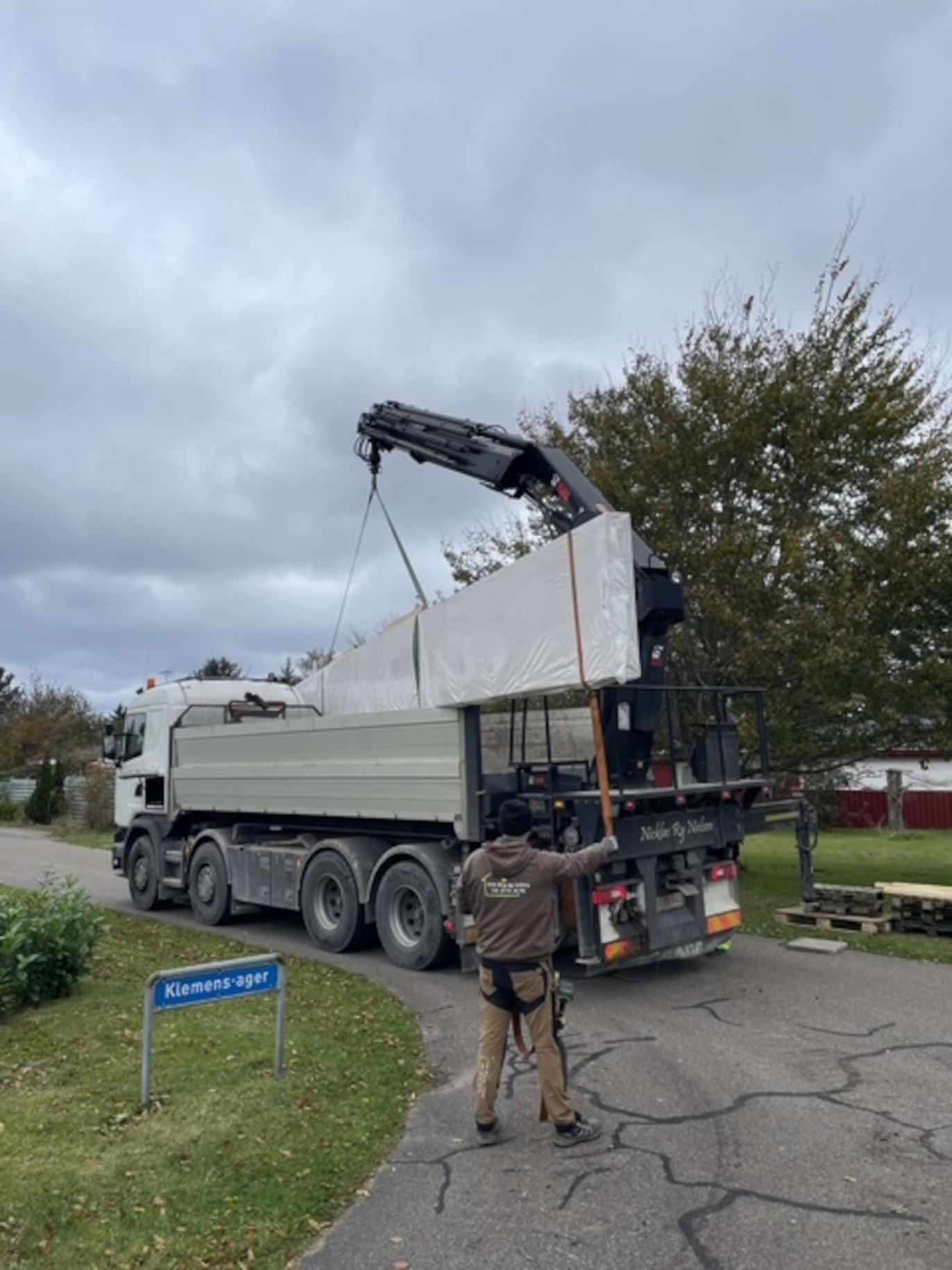 Lastbil med kran løfter en stor metalplade på en vej i et boligområde under en overskyet himmel.