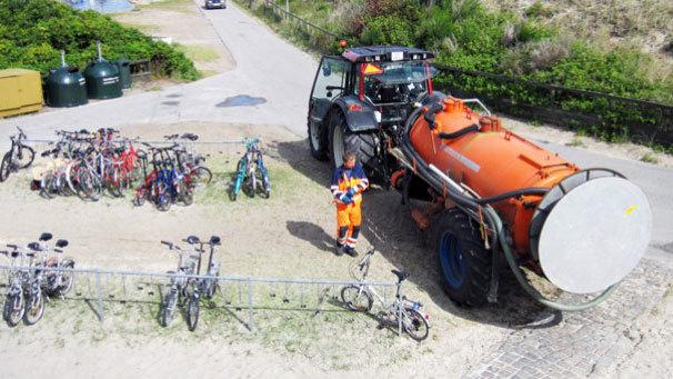 En traktor med en stor tank står ved en cykelparkering, mens en person i arbejdstøj står ved siden af.