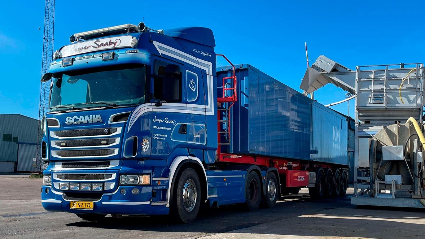 En blå lastbil med en container parkeret ved en havn under en klar blå himmel.