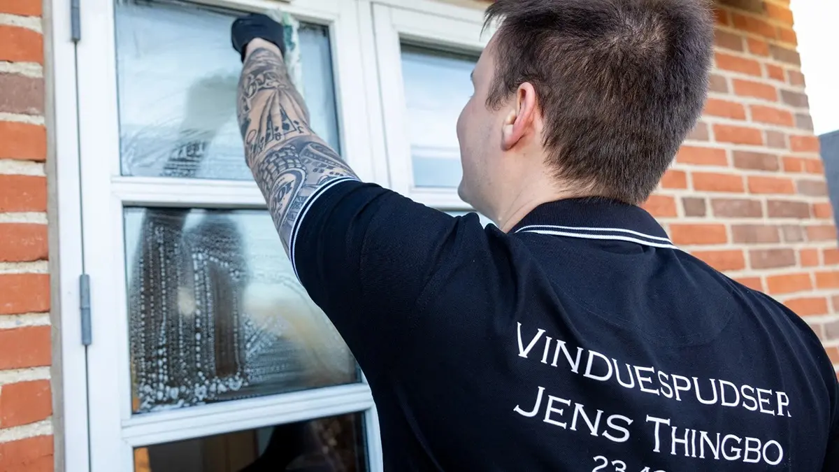 En mand med tatoveringer pudser vinduer på en murstensvæg. Han bærer en sort skjorte med teksten "Vinduespudser Jens Thingbo".