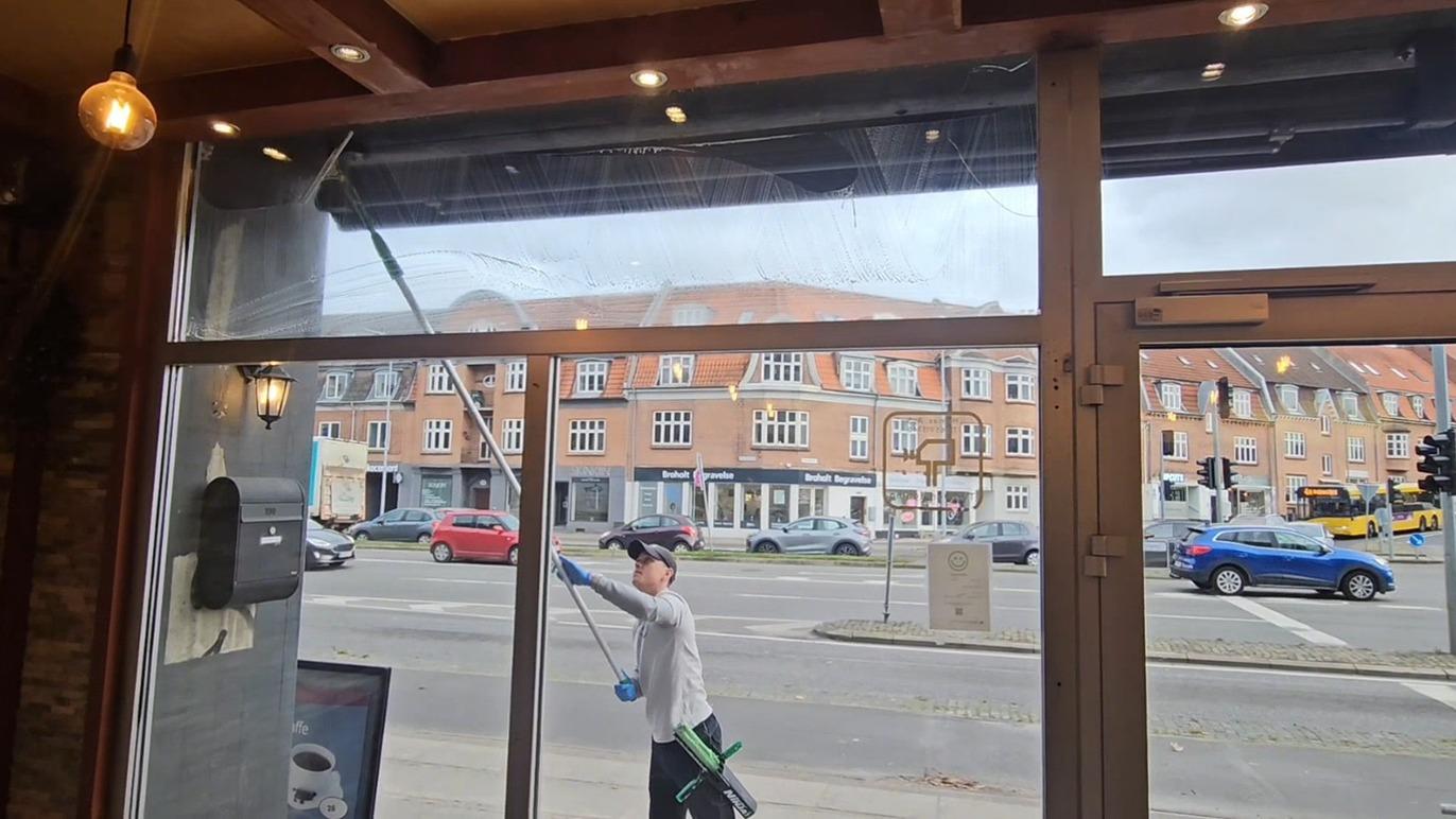 En person vasker vinduet i en café med en lang stang, mens biler kører forbi udenfor.
