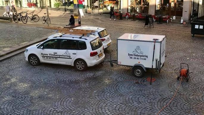 To biler parkeret på en brostensbelagt plads, en hvid bil med tagbøjler og en trailer bagpå.