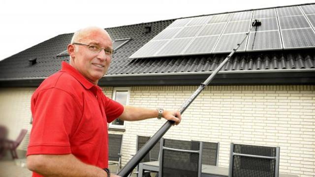 Mand i rød skjorte rengør solpaneler på taget af et hus med en lang stang.