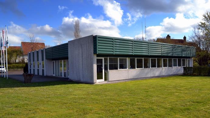 Moderne bygning med grønne tagplader og store vinduer, beliggende på en græsplæne under en skyet himmel.