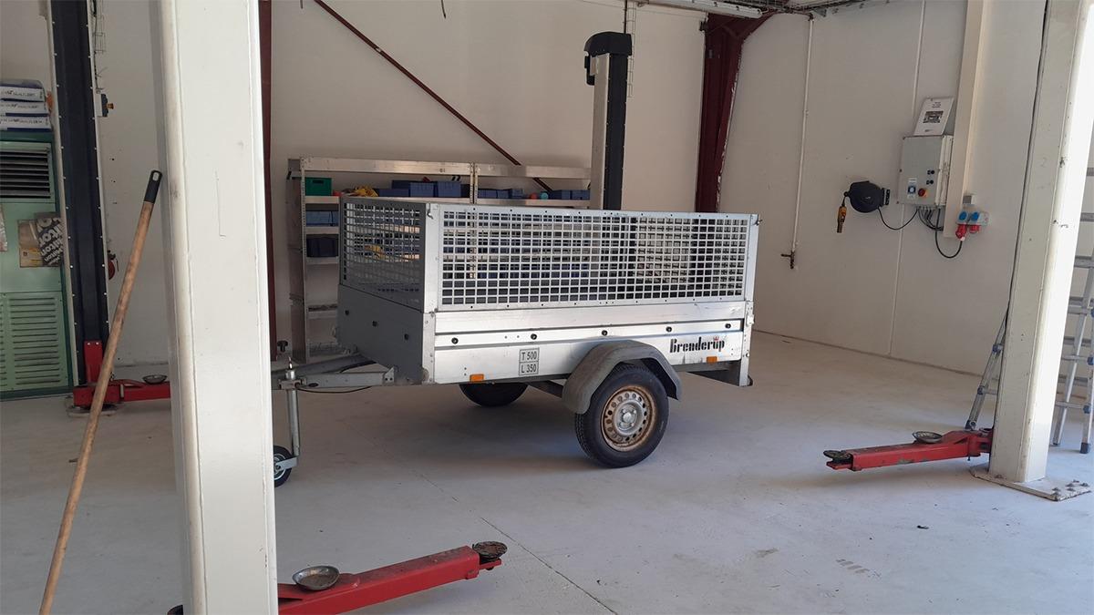En metaltrailer med gitter, parkeret i en garage med værktøj og løfteudstyr i baggrunden.