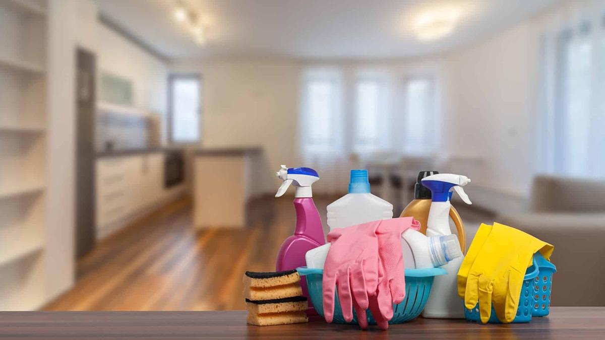 Rengøringsartikler som sprayflasker, svampe og handsker i en kurv på et bord i et lyst køkken.
