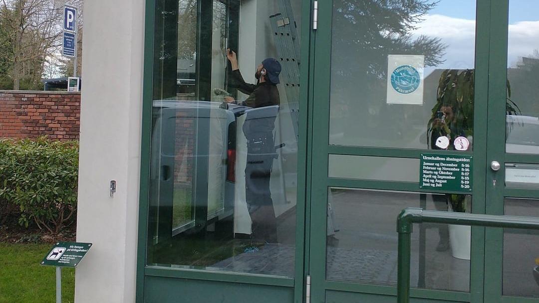 En person rengør vinduet på en glasdør ved en bygning med grønne rammer.