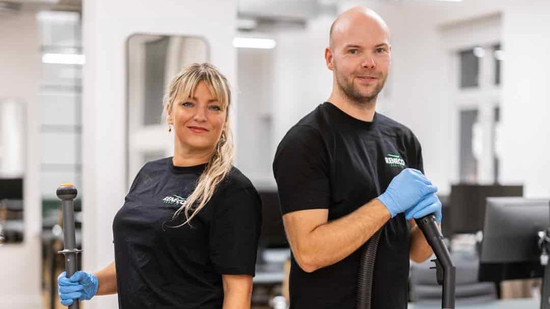 To personer i sorte T-shirts står med rengøringsudstyr i et moderne kontormiljø.