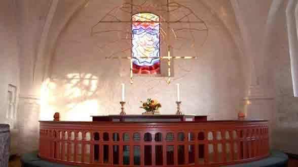 Alteret i en kirke med farvet glasvindue, stearinlys og blomsterdekorationer.