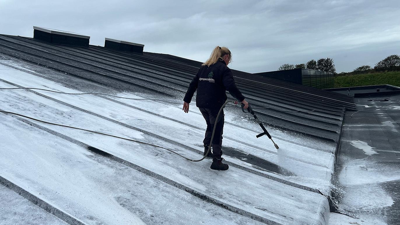 En person står på et tag og rengør det med en højtryksrenser under en grå himmel.