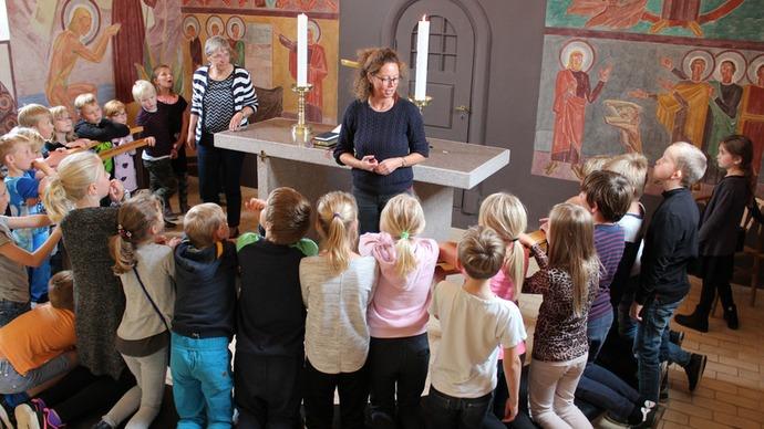 Børn samles omkring en lærer i en kirke med farverige vægmalerier og stearinlys på alteret.
